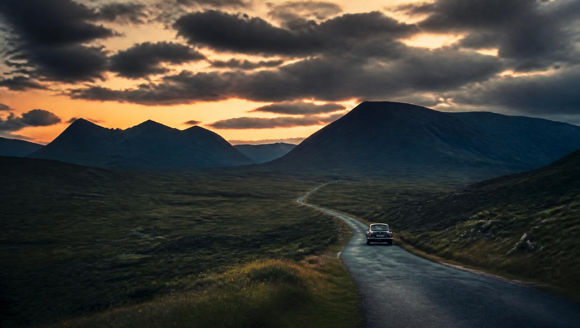 A classic British sports car on a single-track road through the Scottish Highlands at dusk